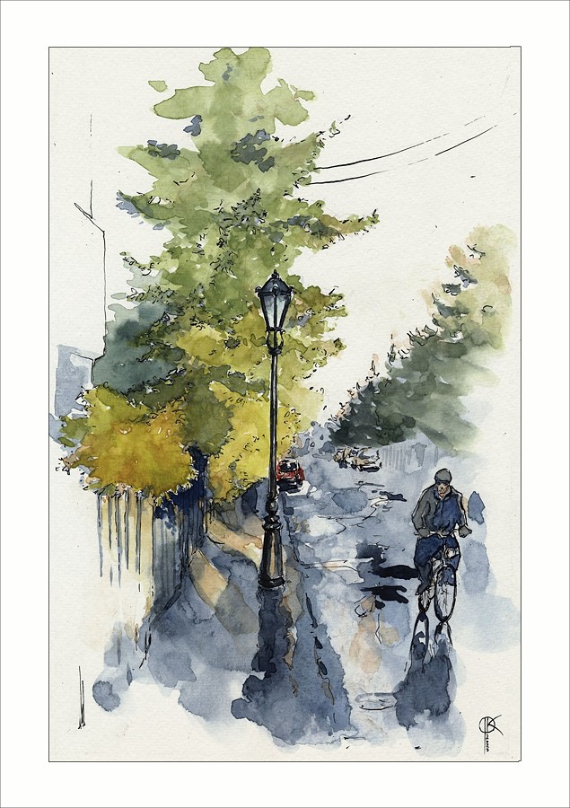 fragment uliczki i chodnika, na chodniku latarnia, w tle drzewa, ulicą jedzie rowerzysta